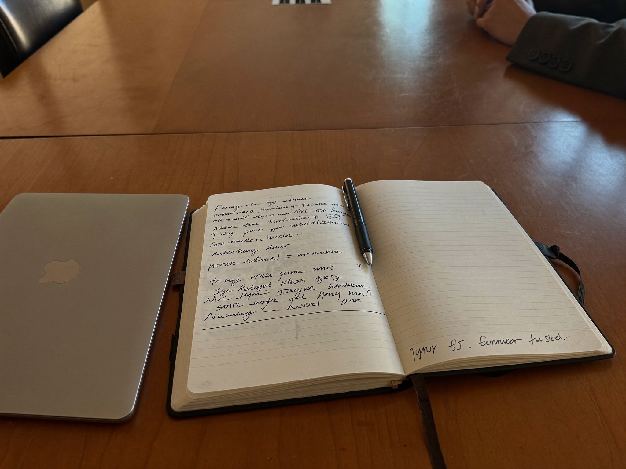 Ahşap bir masada el yazısıyla dolu açık bir defter, yanında bir toplantı transkripti gösteren telefon